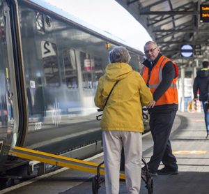 Staff member assisting passenger with mobility aid onto train with ramp deployed at a train station