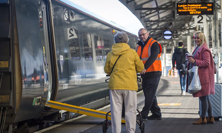 Staff member assisting passenger with mobility aid onto train with ramp deployed at a train station