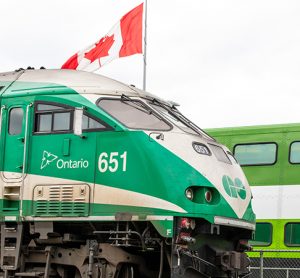 Torontobound Metrolinx GO transit MotivePower Industries Corporation locomotive 651 pulling a commuter train through a station past a Canadian flag.