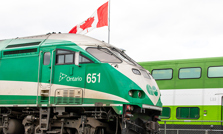 Torontobound Metrolinx GO transit MotivePower Industries Corporation locomotive 651 pulling a commuter train through a station past a Canadian flag.